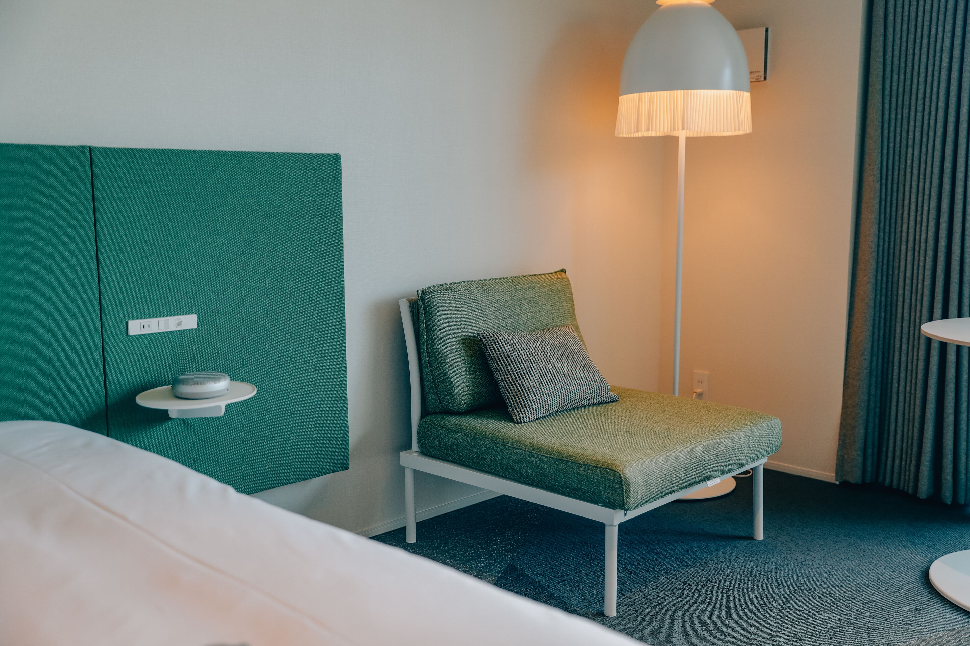 Una habitación de hotel con cama, un sillón verde tapizado con un cojín, una mesita de noche con lámpara y un panel divisor verde.