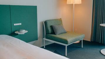 Una habitación de hotel con cama, un sillón verde tapizado con un cojín, una mesita de noche con lámpara y un panel divisor verde.
