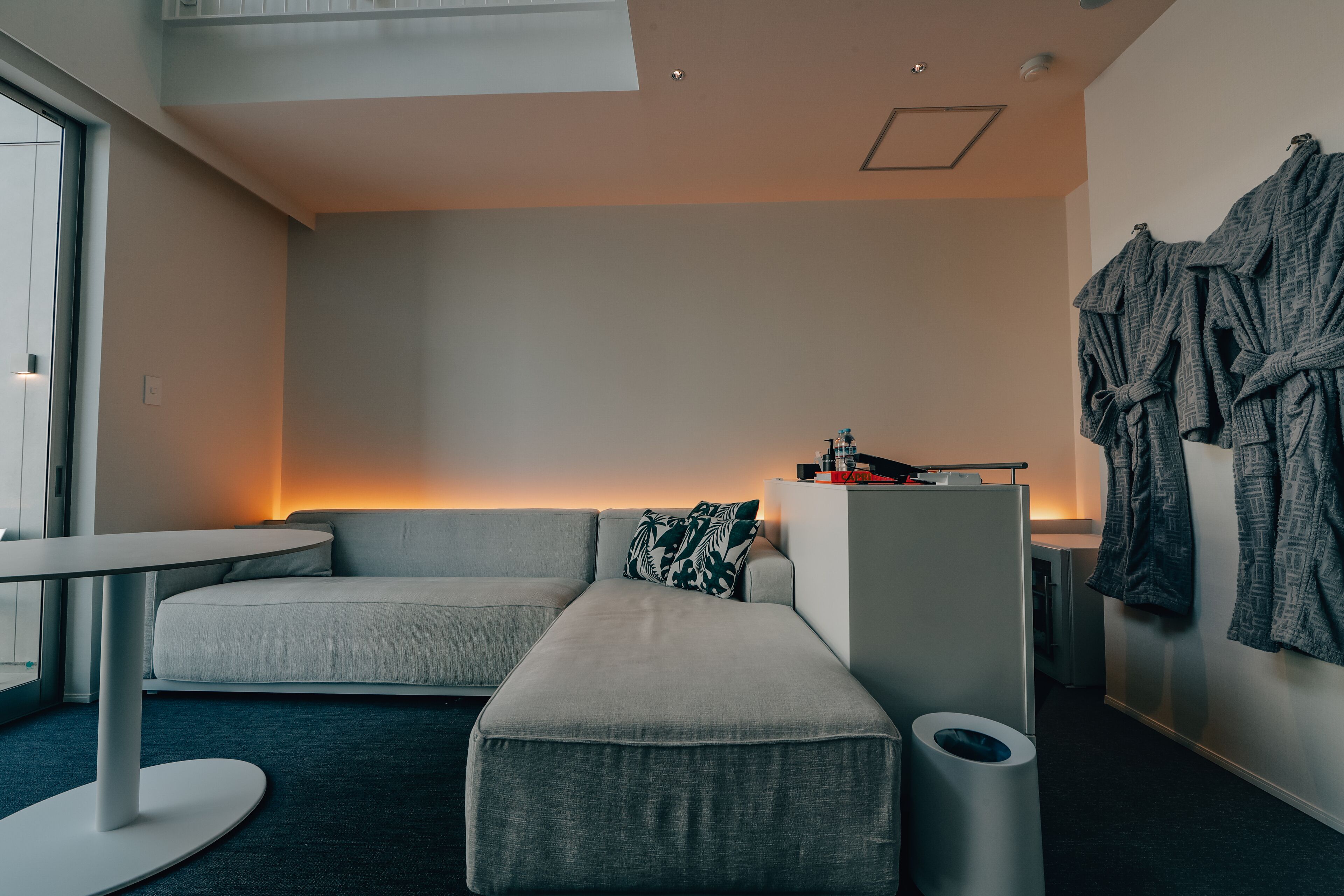 Moderni hotellihuone, jossa on harmaa sohva, pieni pöytä ja penkki. Seinällä on henkarilla aamutakkeja ja penkin lähellä on roskakori.