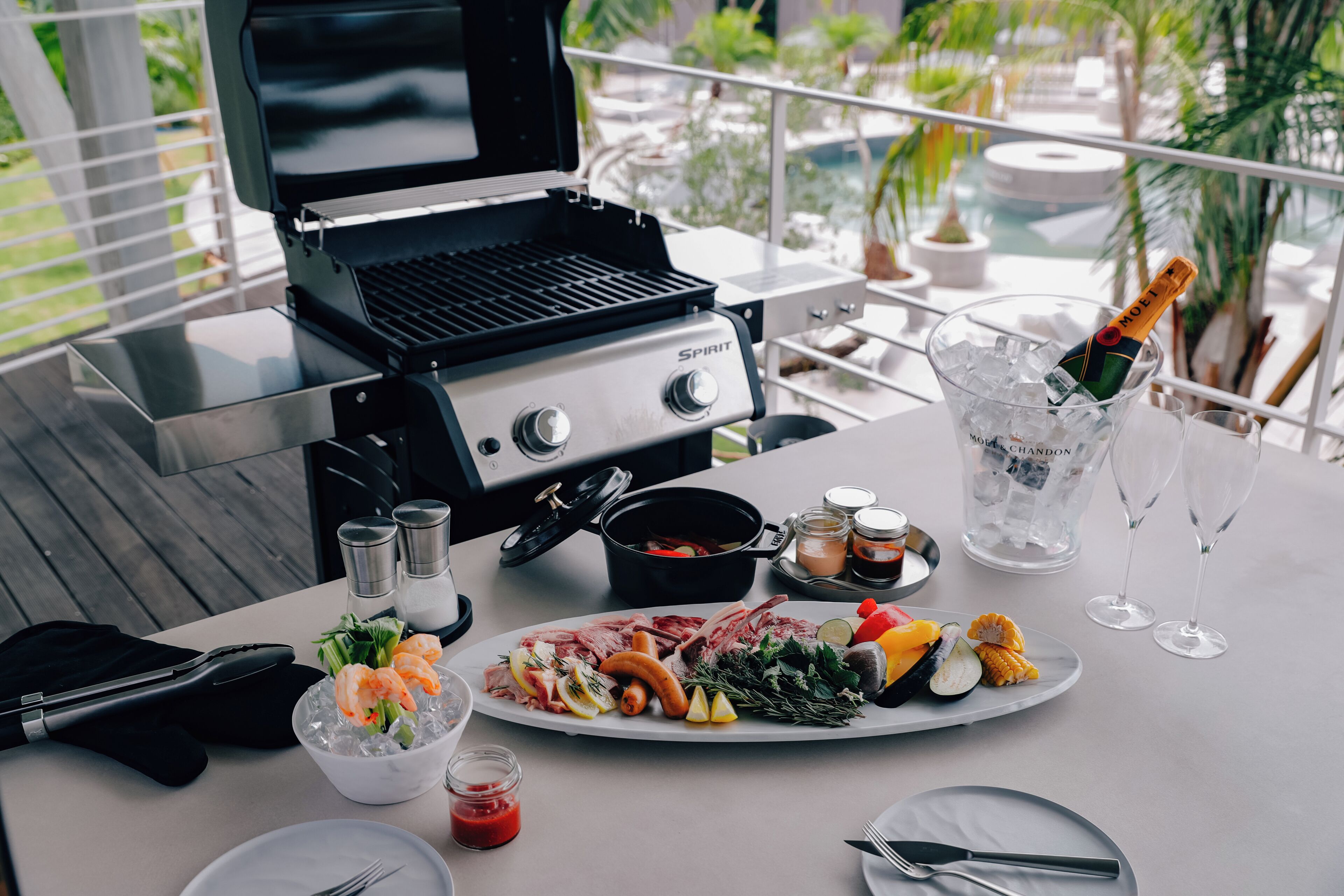 A barbecue grill, a variety of meats, vegetables, and a bottle of champagne on an outdoor table.