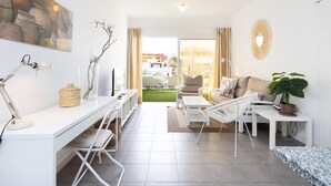 En moderne stue med et hvidt skrivebord, en hvid sofa og et spisebord. Et stort vindue med gardiner, en plante og udsigt udendørs.