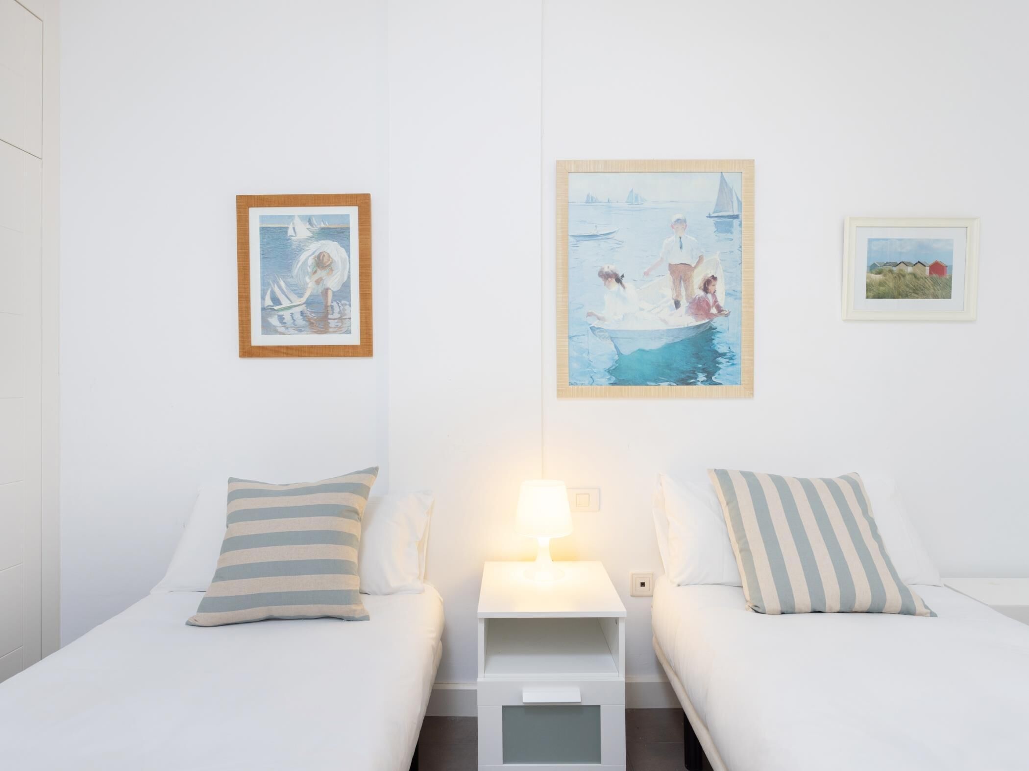 A hotel room with two beds, a nightstand with a lamp, and three paintings on the wall.
