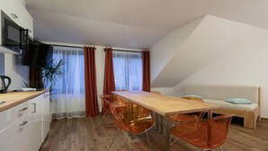 A modern kitchen with a wooden countertop, a built-in microwave, and a sink. There is a dining area with a wooden table and four orange chairs. A sofa with a light blue cushion is against the wall, and a potted plant is on the countertop.