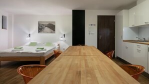 A modern hotel room with a wooden dining table, a bed with green pillows, a black door, and a kitchen area with white cabinets.
