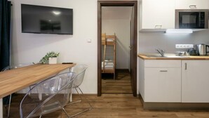 Een kleine keuken met witte kasten, een magnetron en een spoelbak. Een houten tafel met stoelen staat op de voorgrond. Een televisie is aan de muur bevestigd, en een stapelbed is zichtbaar op de achtergrond.