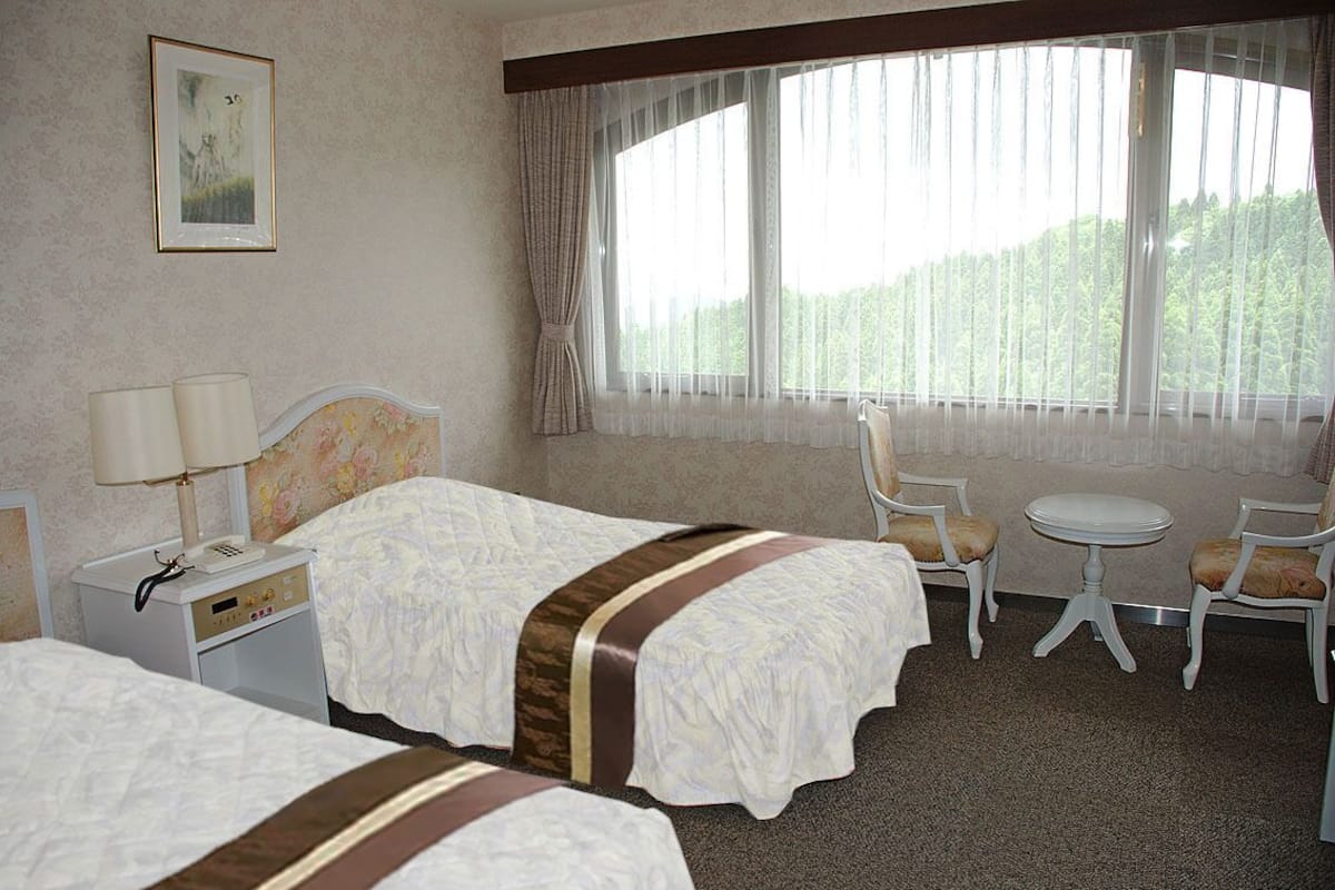A hotel room with two beds, a small table, two chairs, a nightstand with a lamp, and a large window with curtains.