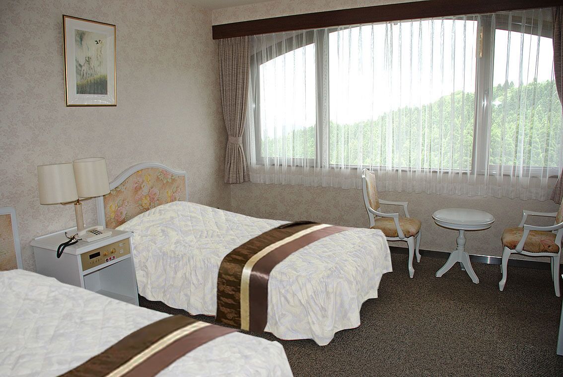 A hotel room with two beds, a desk with a lamp, a chair, a small table, and a large window with curtains.