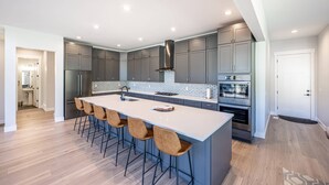 A modern kitchen with grey cabinets, a central island with bar stools, stainless steel appliances, and a tiled backsplash.