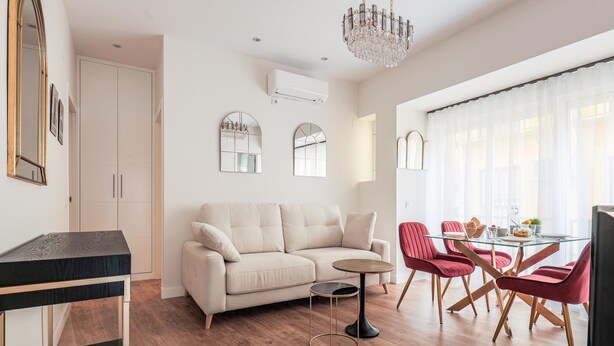 A modern living room with a beige sofa, a glass dining table with red chairs, a chandelier, and a large window with sheer curtains. - Pure Seville elegance and comfort (Sevilla)