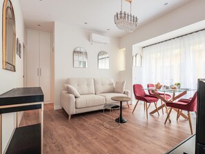 Een moderne woonkamer met een beige bank, een glazen eettafel met rode stoelen, een kroonluchter en een groot raam met dunne gordijnen.