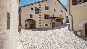 Una calle empedrada que conduce a un edificio con fachada tradicional, contraventanas de madera y un pequeño balcón con flores.
