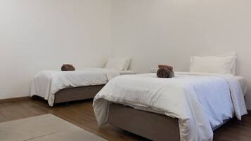Una habitación con dos camas, cada una con ropa blanca y una almohada marrón. La habitación tiene suelo de madera y paredes blancas.