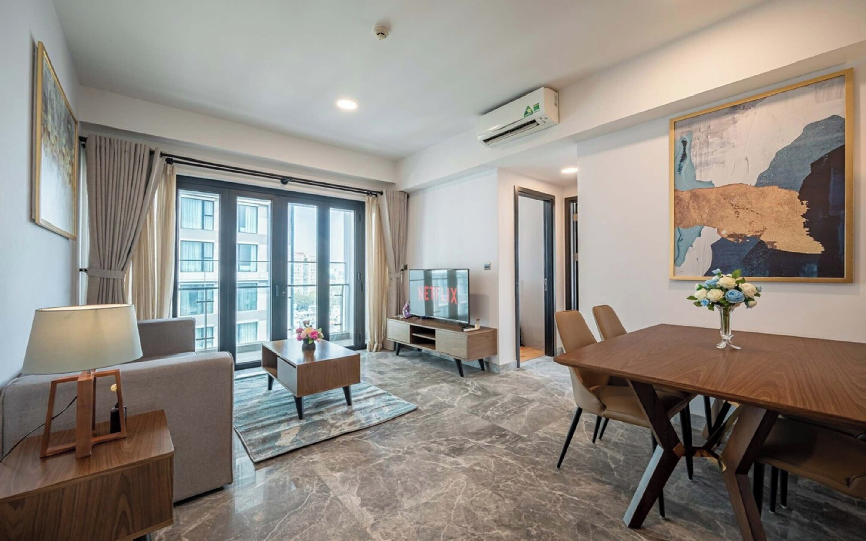 A modern living room with a sofa, a coffee table, a TV, and a dining table with chairs. There is a large window with curtains, an air conditioning unit, and a painting on the wall.