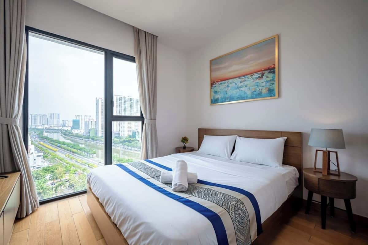 A hotel room with a large window offering a city view, a neatly made bed, a painting on the wall, and a bedside table with a lamp.