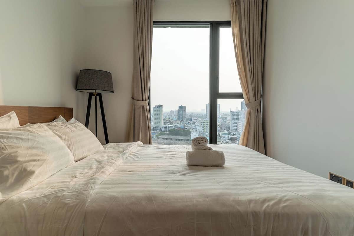 A hotel room with a large bed, a lamp, and a view of a cityscape through a window with curtains.