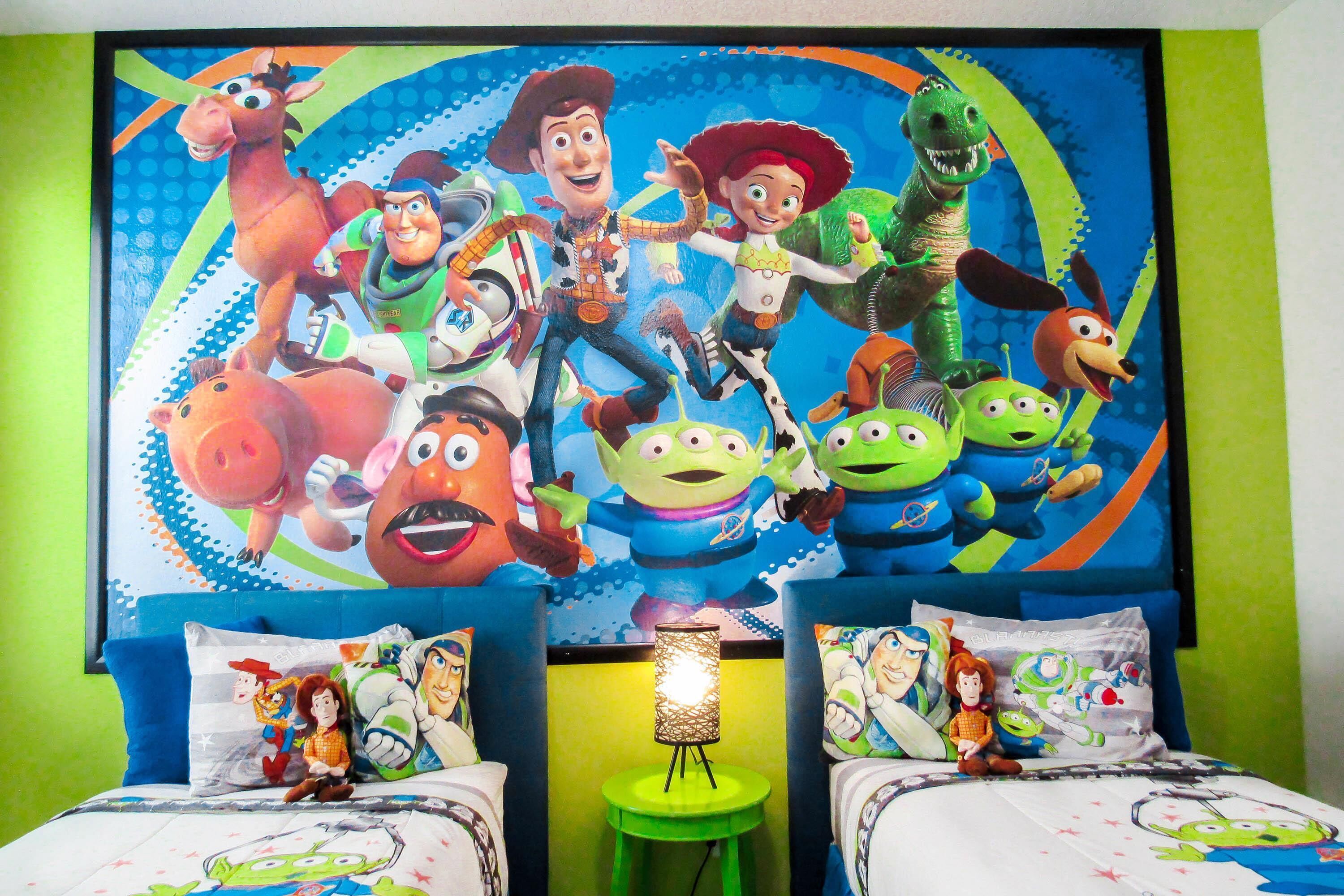 Una habitación infantil con un mural grande de un personaje animado en la pared, dos camas con ropa de cama a juego, y un taburete verde pequeño con una lámpara.