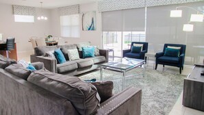 A spacious living room with a grey sofa set, a glass coffee table, and a dining area with a table and chairs. - Spacious 6 BR Home with Pool, Spa, Lanai, Grill, Wet Bar in Solara Resort! (Kissimmee)