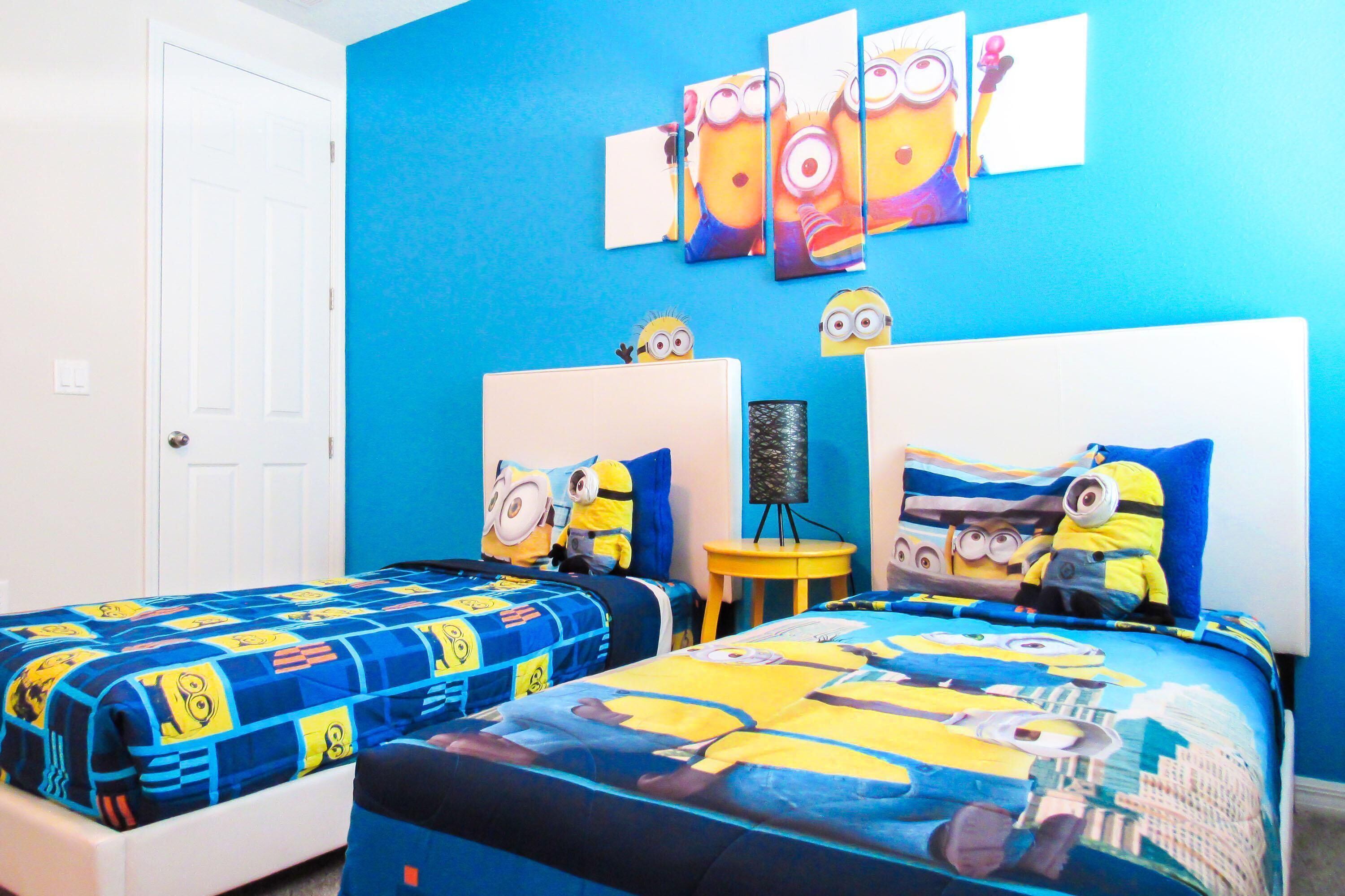 A bedroom with two beds, each covered with a Minion-themed duvet. There are wall art pieces and a small table with a lamp.