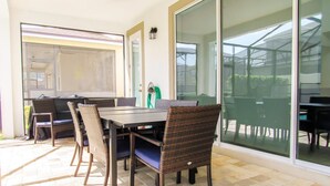 A covered patio with a dining table and chairs, a green hose, and a sliding glass door leading to a garden. - Spacious 6 BR Home with Pool, Spa, Lanai, Grill, Wet Bar in Solara Resort! (Kissimmee)