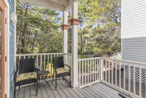 A covered porch with two wicker chairs and a small table, surrounded by white railings and a view of a wooded area.