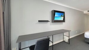 Habitación de hotel con un escritorio gris, un televisor montado en la pared mostrando una escena de playa y una cama con cabecera blanca.