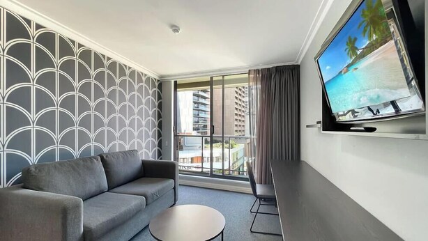 Habitación de hotel moderna con pared de diseño gris, un sofá gris, una mesa de centro redonda, un televisor en la pared y un balcón con vistas a palmeras y una playa.