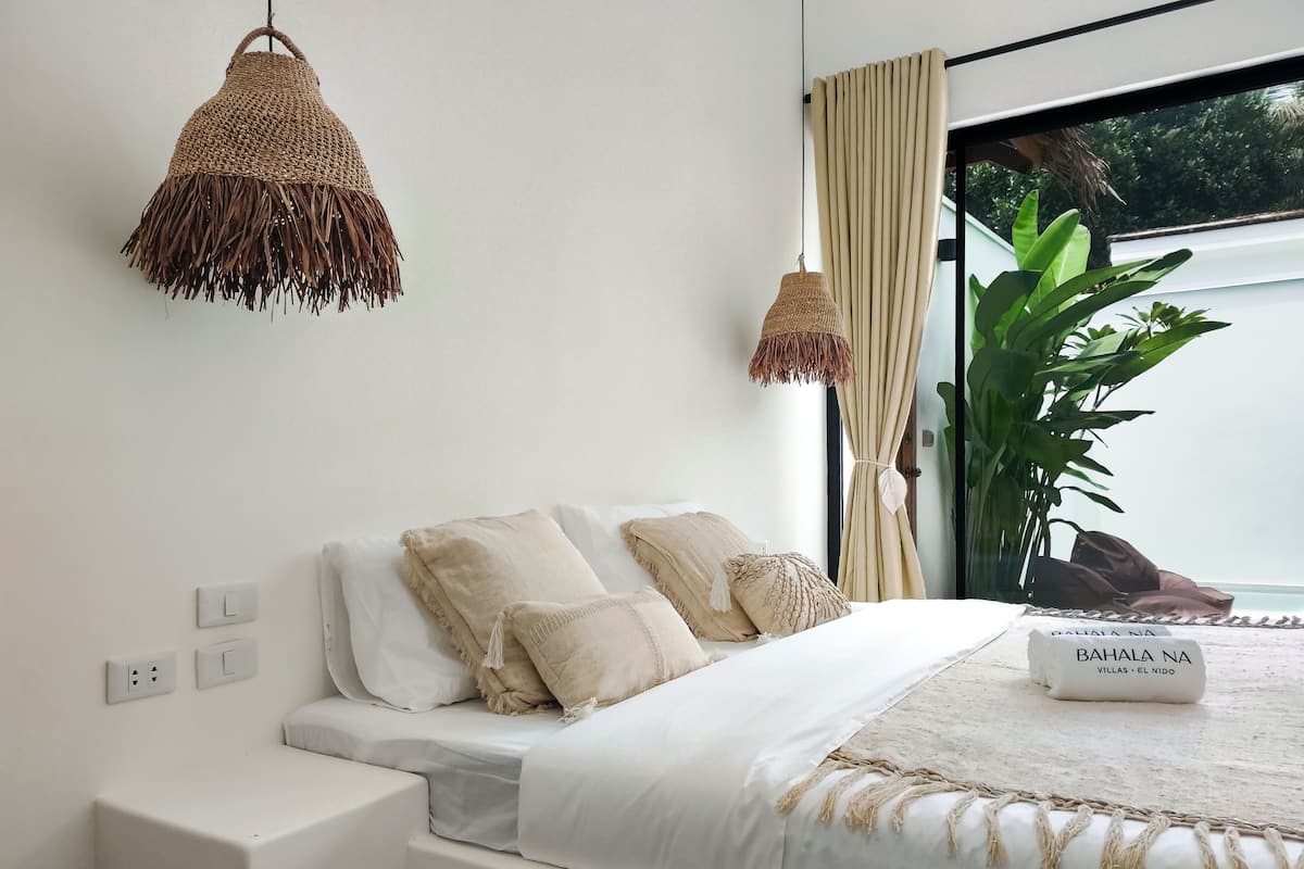 A modern bedroom with a large bed, two hanging lamps, a window with curtains, and a plant.