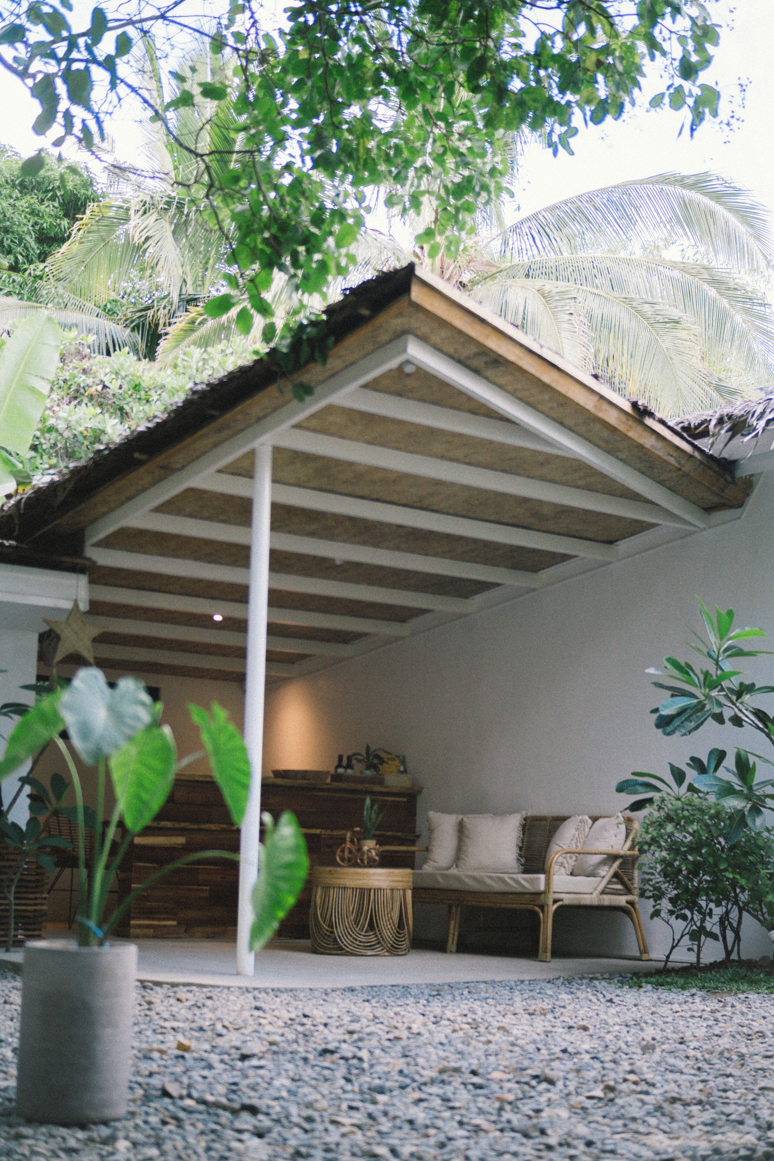 A covered outdoor seating area with a wooden bench and a woven coffee table, surrounded by potted plants and a gravel floor.