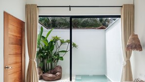 A modern room with a wooden door, beige curtains, a large window with a view of palm trees, a bed with a white towel, and a hanging lamp. - Bahala Na Villas (El Nido)