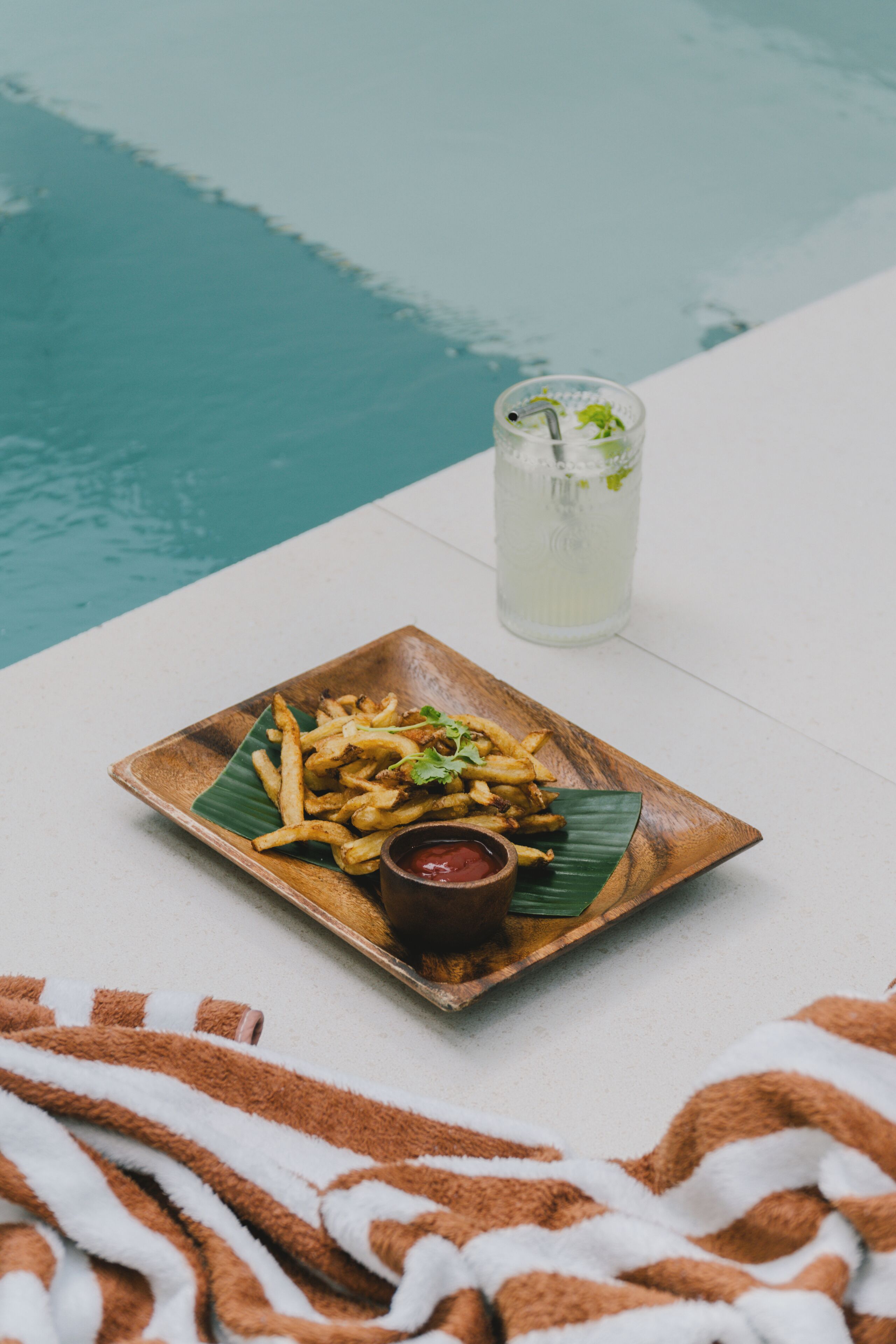 Ein Holzteller mit Pommes und einer kleinen Ketchup-Schale, ein Glas Eistee sowie ein Bananenblatt auf einer weißen Fläche in der Nähe eines Pools.
