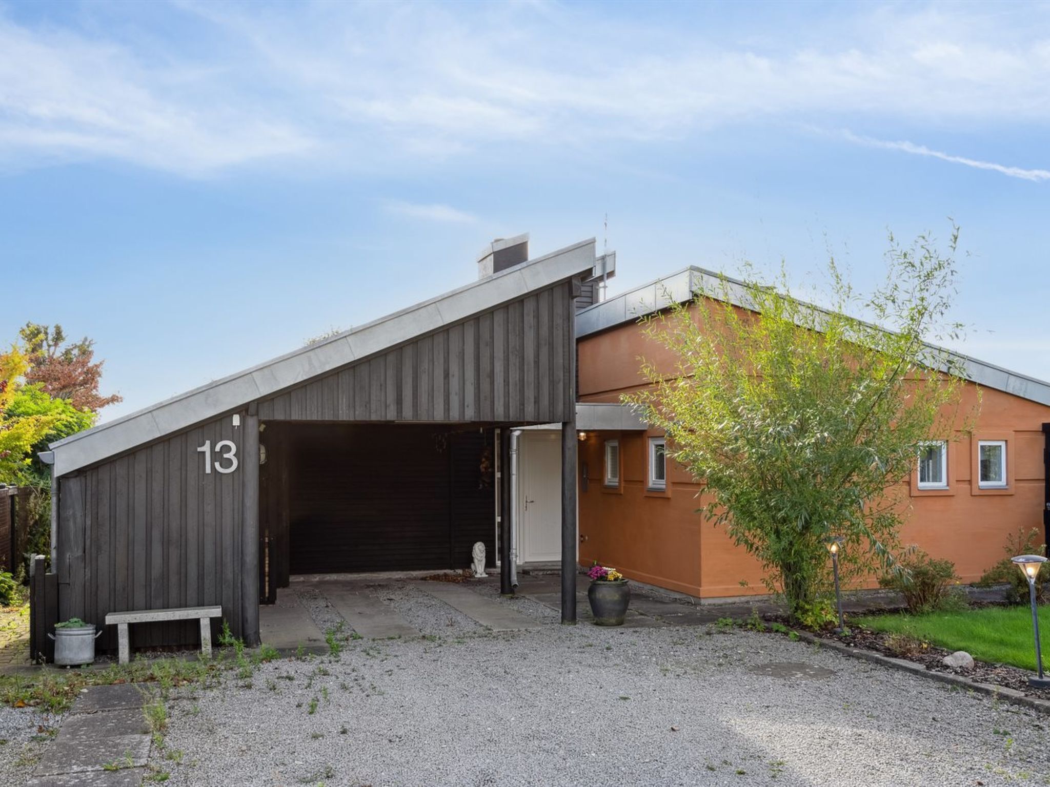 Um edifício moderno com entrada de garagem, uma placa com o número 13 e uma pequena área de jardim com um banco e plantas em vasos.