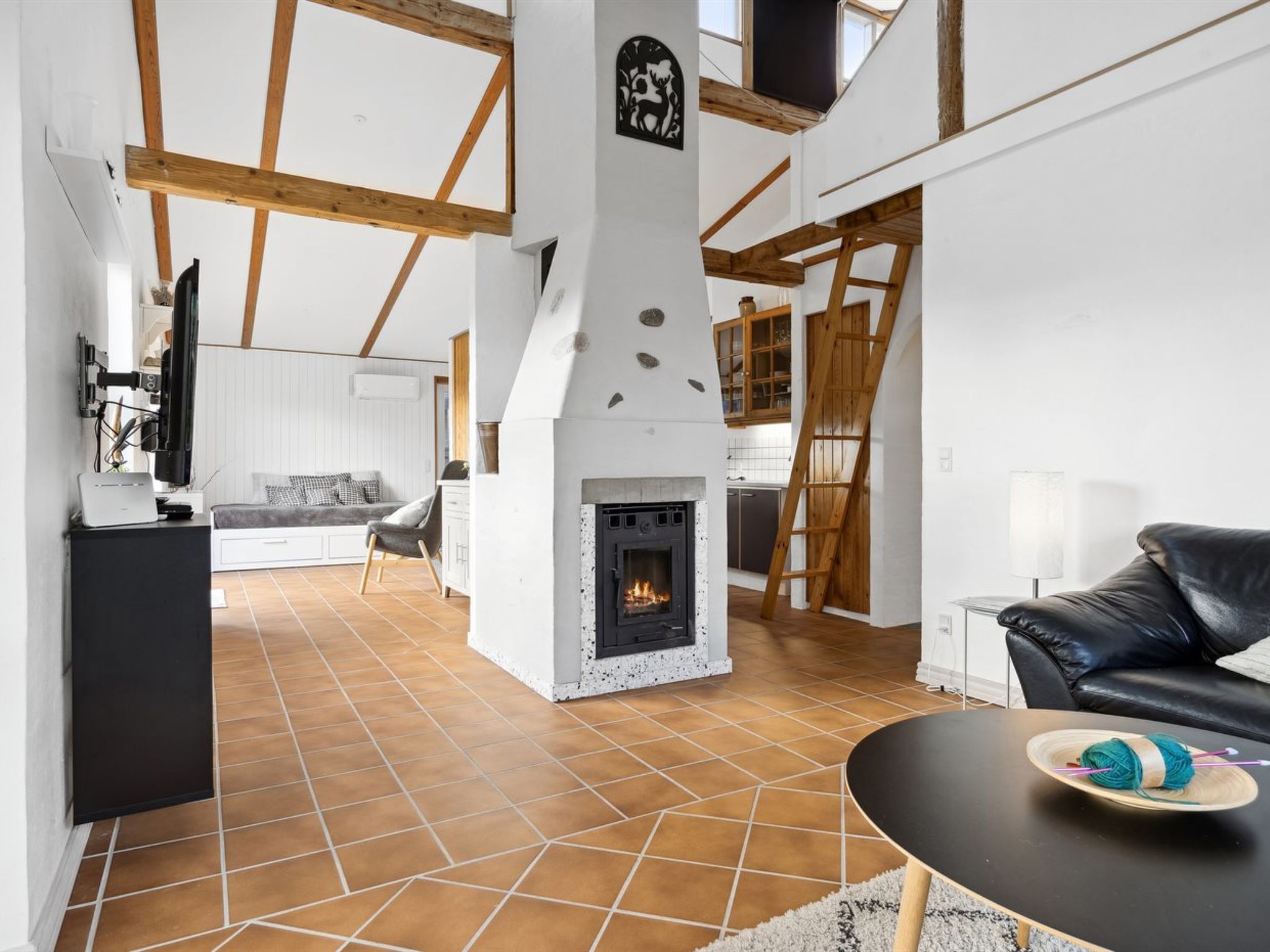 A spacious living area with a fireplace, wooden beams, and a kitchen in the background.