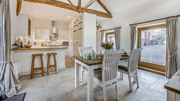 A kitchen with white cabinets, a wooden island, and stools. There is a dining table with chairs and a window with curtains. - Two bedroom barn conversion in the Cotswolds - The Byres (Elmley Castle, Pershore)