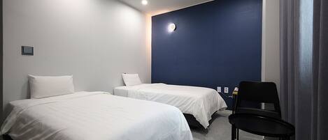 A hotel room with two single beds, a chair, a desk with a computer, and a blue accent wall.