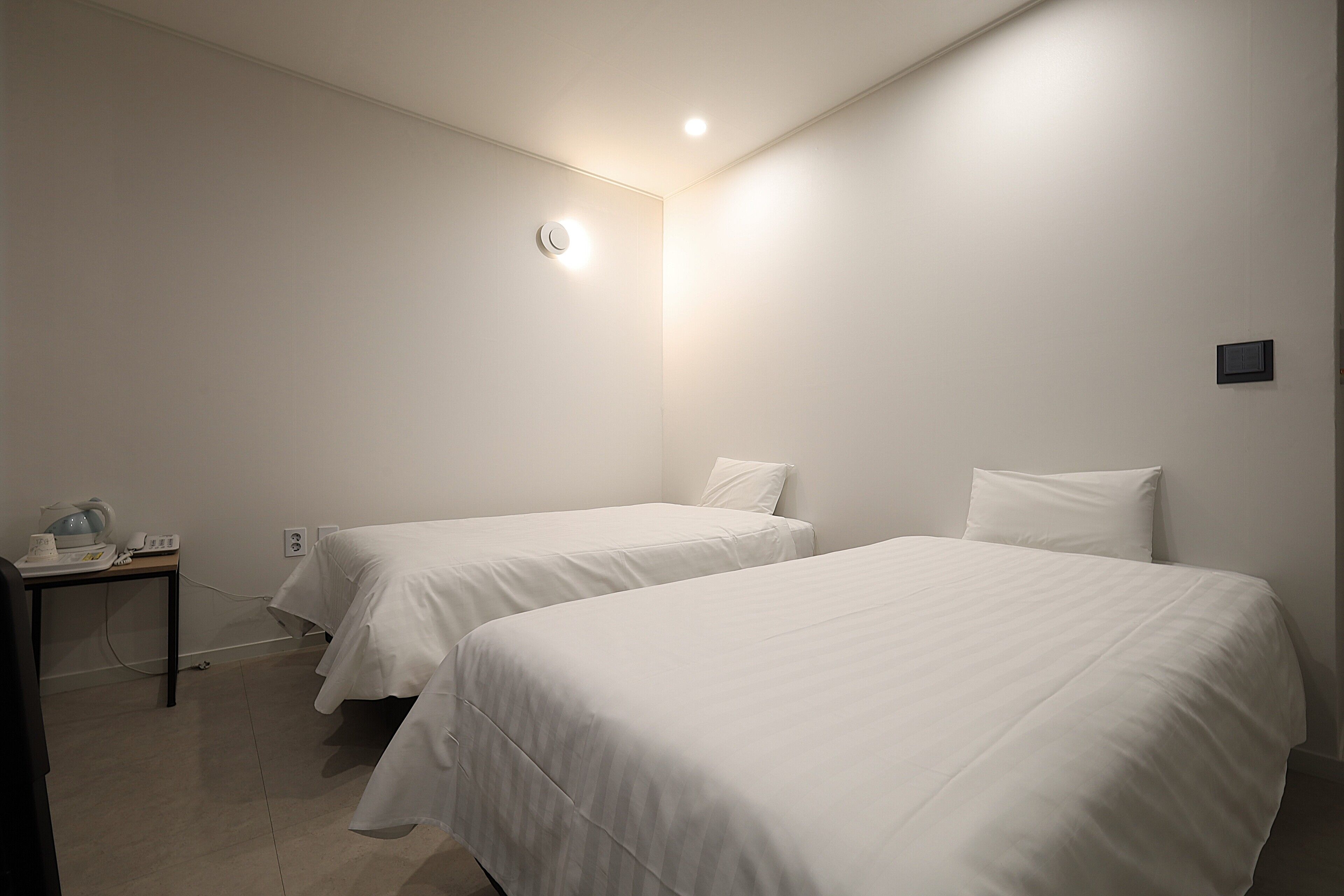 A hotel room with two single beds, a small table with a lamp, and a wall-mounted light fixture.