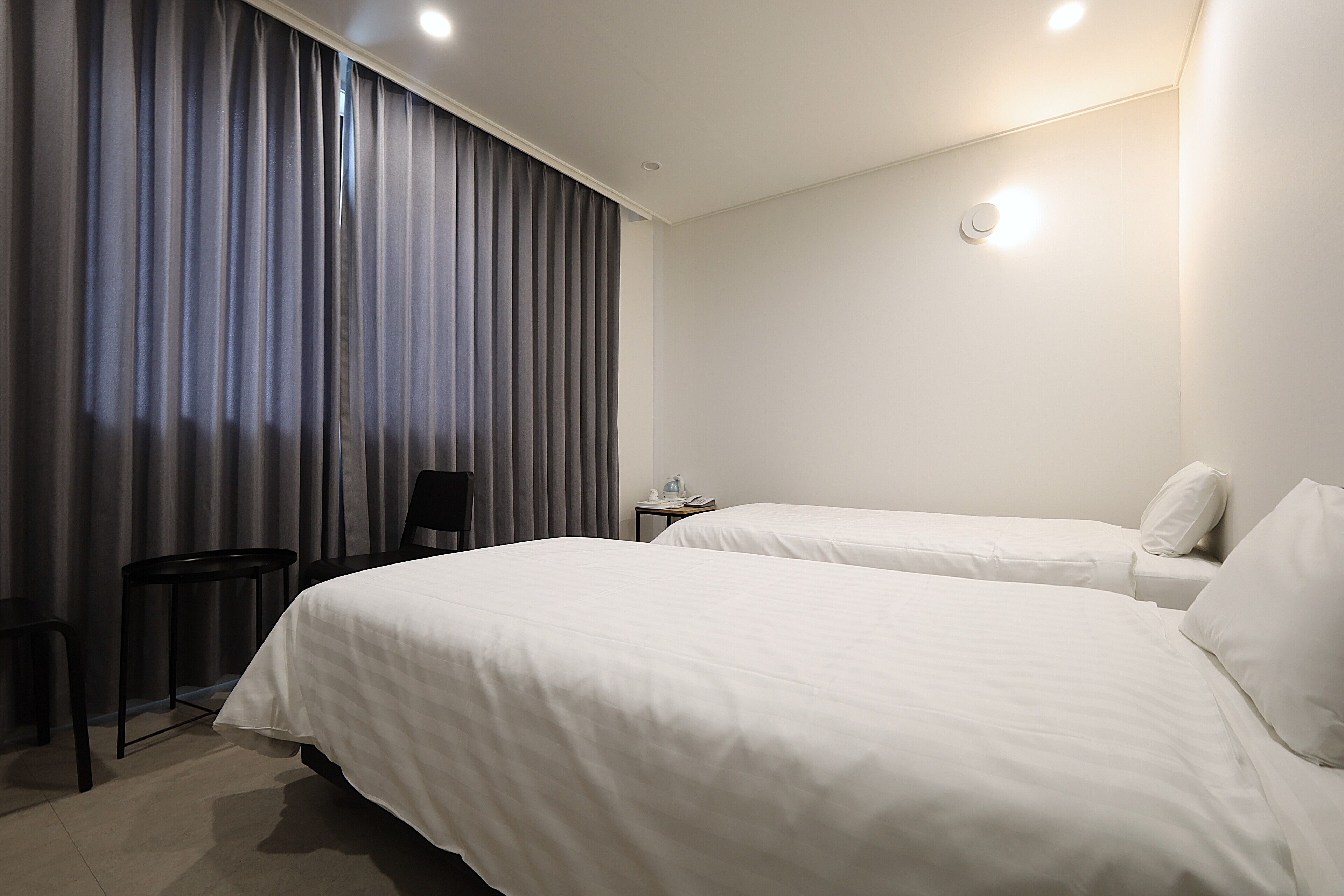 A hotel room with two beds, a small table, a chair, and a window with curtains.