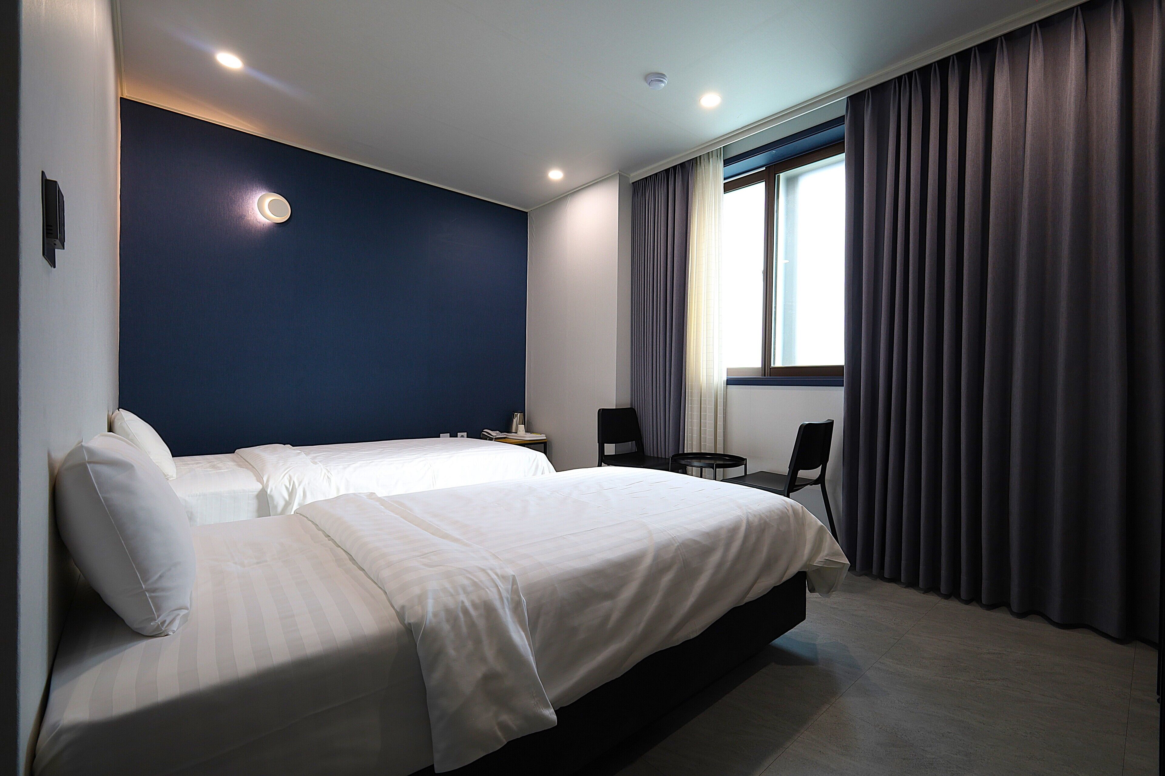 A hotel room with two beds, a small table, a chair, and a window with curtains.