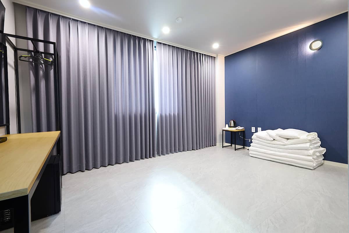 Une chambre d’hôtel avec une grande fenêtre ornée de rideaux gris, un bureau en bois, une petite table avec une lampe et une pile de serviettes blanches adossées à un mur bleu.