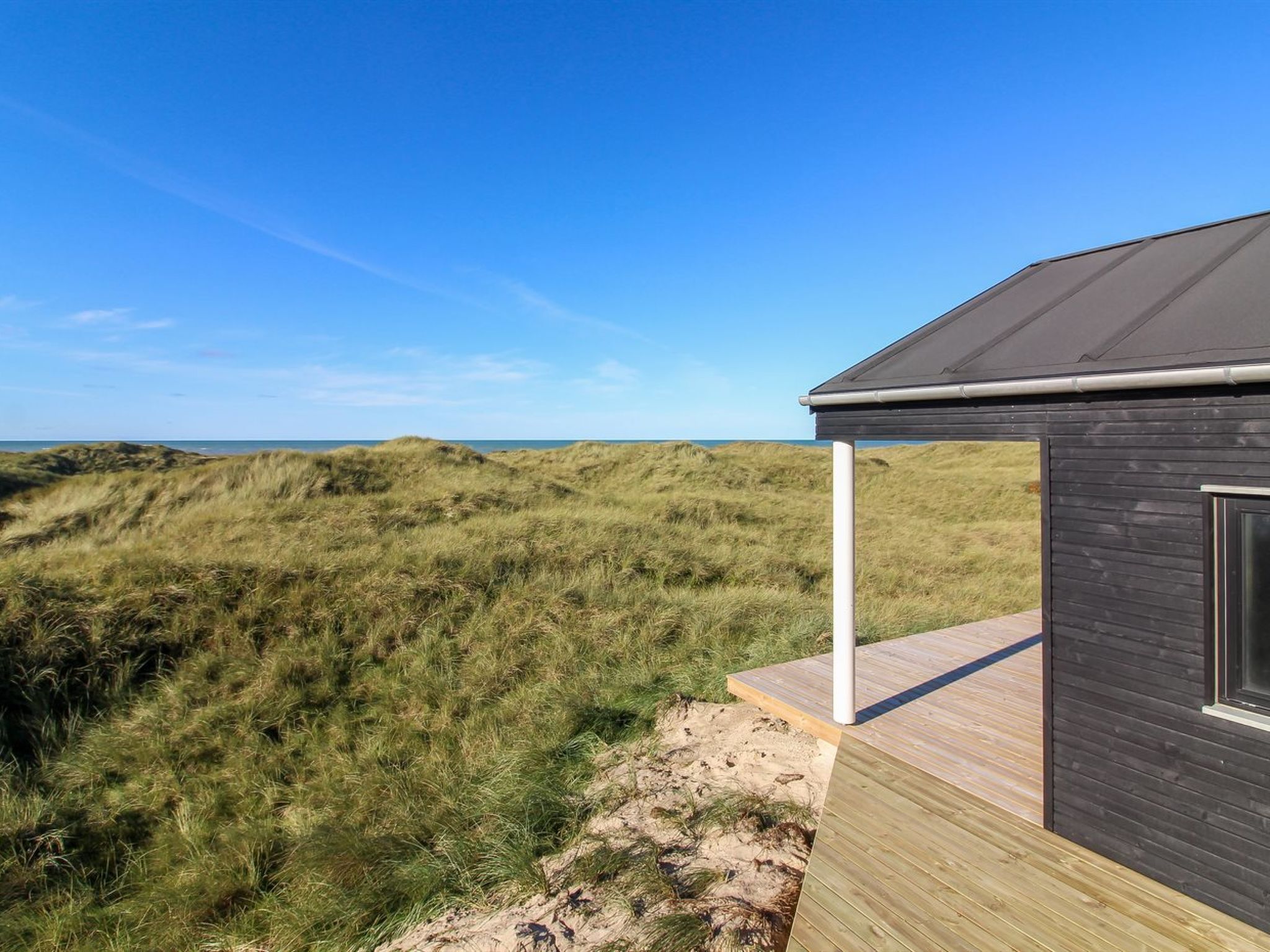 A modern building with a dark roof and a wooden deck overlooking a grassy dune landscape.