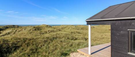 A modern building with a dark roof and a wooden deck overlooking a grassy dune landscape.