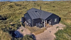 Uma casa moderna preta com um deck, situada numa praia arenosa com dunas e o oceano ao fundo.