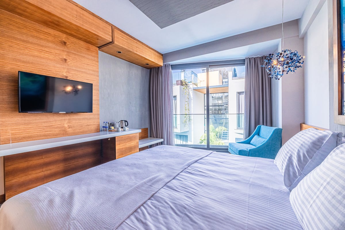 Habitación de hotel moderna con una cama grande, un televisor de pantalla plana en la pared, un rincón de descanso con una silla azul y un ventanal con vistas a la ciudad.