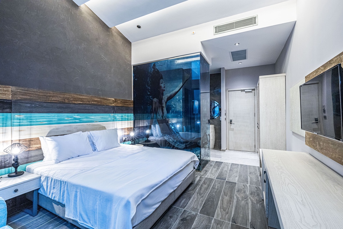 A modern hotel room with a large bed, a glass wall, and a bathroom with a mirror and sink.