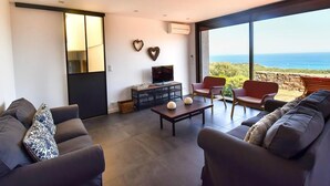 A modern living room with a large window, a flat-screen TV, and a sofa. - Villa in Gaou with Sea View and Beach (Bormes-les-Mimosas)