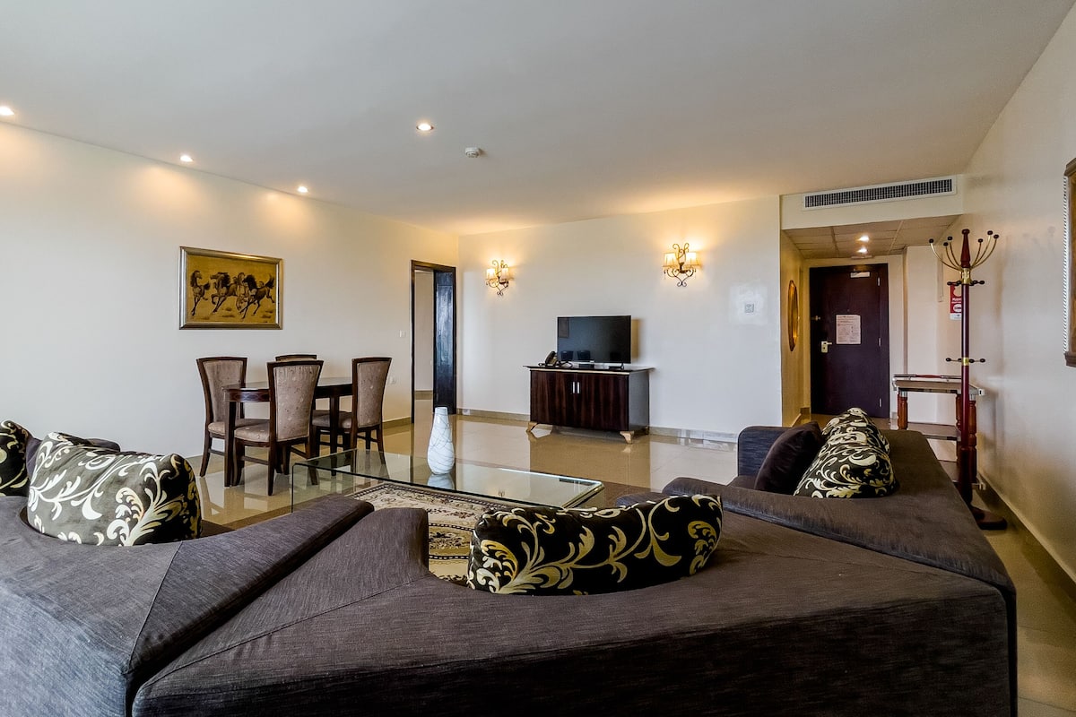 A living room with a glass coffee table, dark sofas with patterned cushions, a dining area with chairs, a TV on a cabinet, and wall-mounted lights.