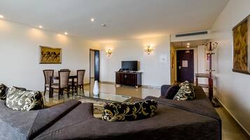 A living room with a glass coffee table, dark sofas with patterned cushions, a dining area with chairs, a TV on a cabinet, and wall-mounted lights.