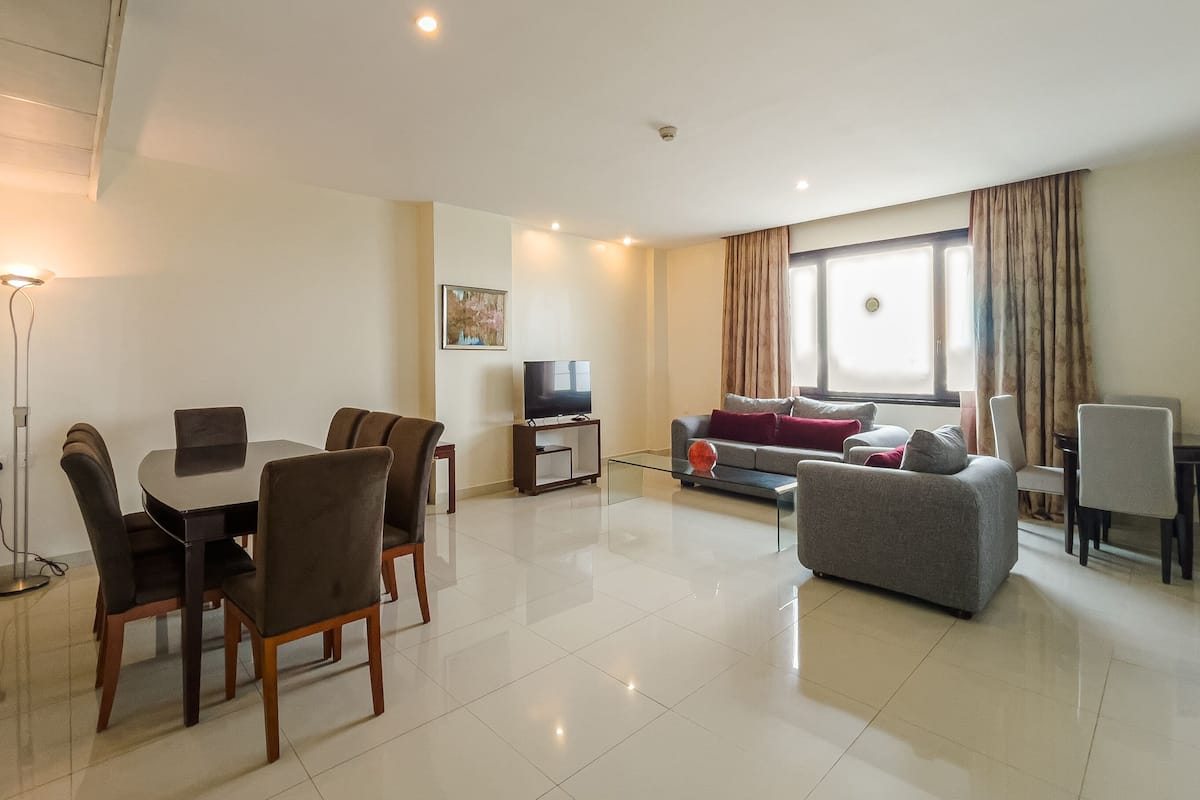 A modern living room with a dining area, a flat-screen TV, and a large window with curtains.