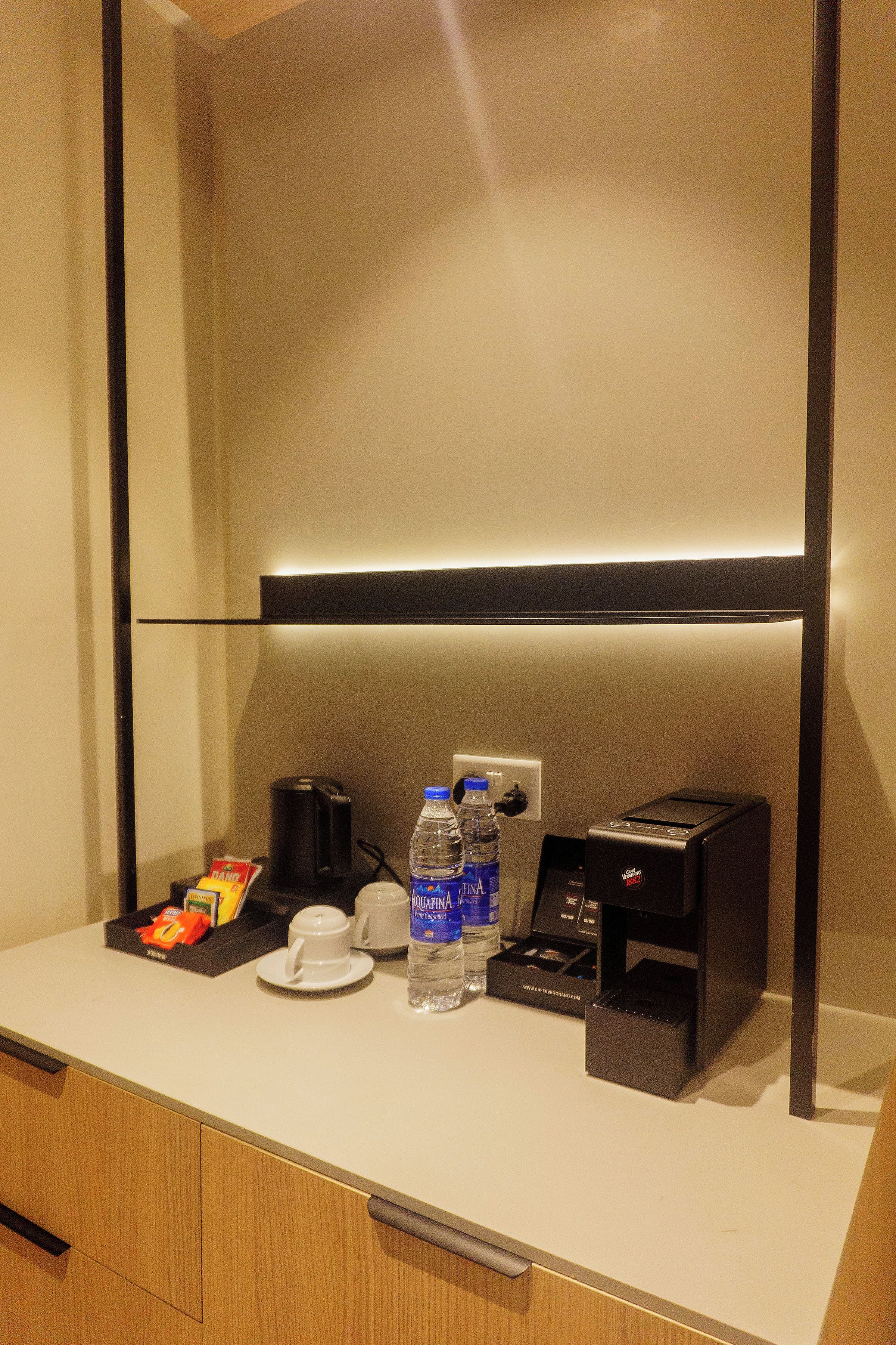 Habitación de hotel con minibar, cafetera, botella de agua, taza y una pequeña caja, probablemente con artículos de tocador.