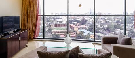 A modern living room with a large window, a flat-screen TV, a sofa with cushions, and a glass coffee table.