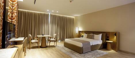 A hotel room with a large bed, a desk with chairs, a small table, and a large window with curtains.